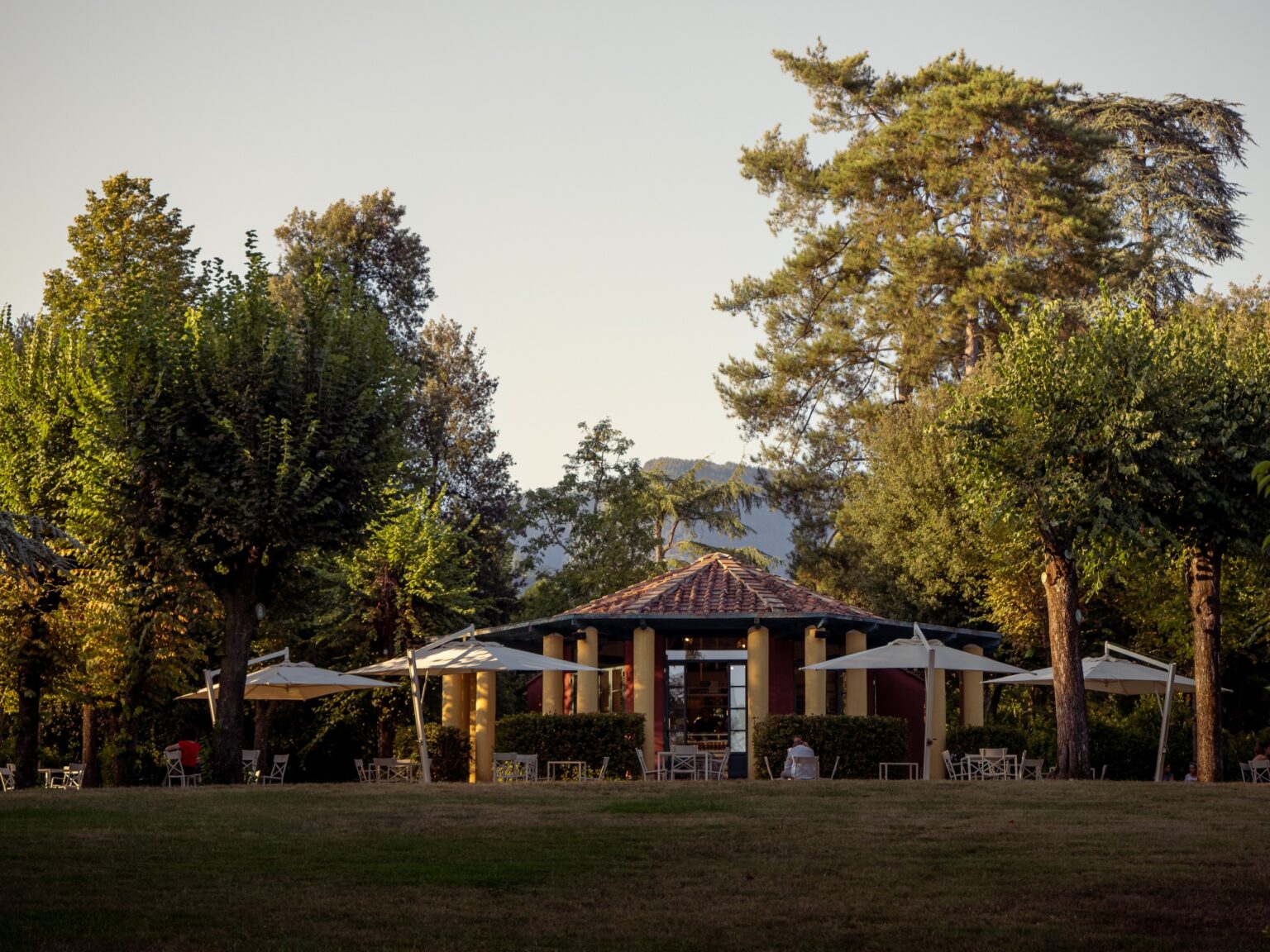 Scenic view of a charming outdoor café surrounded by lush trees and patio umbrellas, set against a backdrop of mountains and a clear sky. Ideal for relaxation and dining in a natural setting. | Only on SopranoVillas