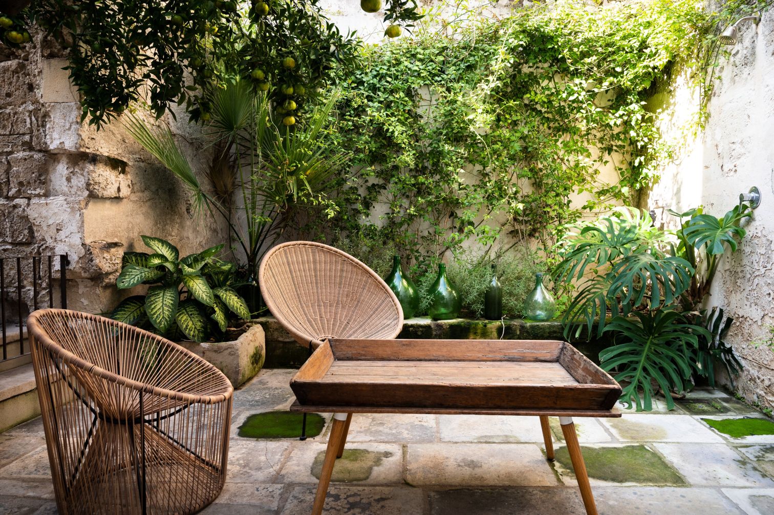 Cozy outdoor garden space featuring two stylish woven chairs, a wooden tray table, and lush greenery with various plants and decorative glass bottles. The setting exudes tranquility and natural beauty, perfect for relaxation and outdoor gatherings. | Only on SopranoVillas