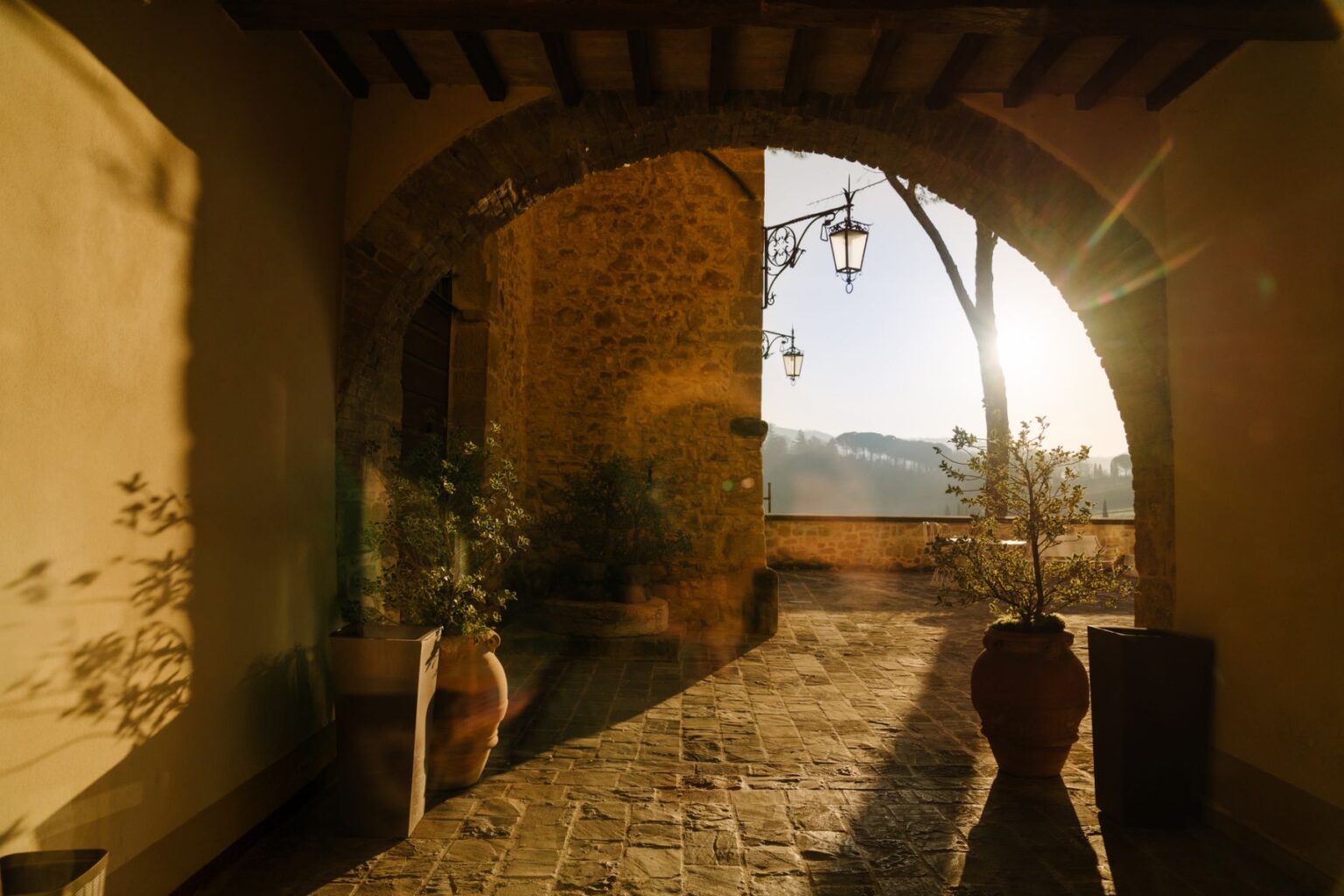Sunlit archway with stone walls and hanging lanterns, featuring potted plants on a cobblestone floor, creating a serene atmosphere in a rustic setting. | Only on SopranoVillas