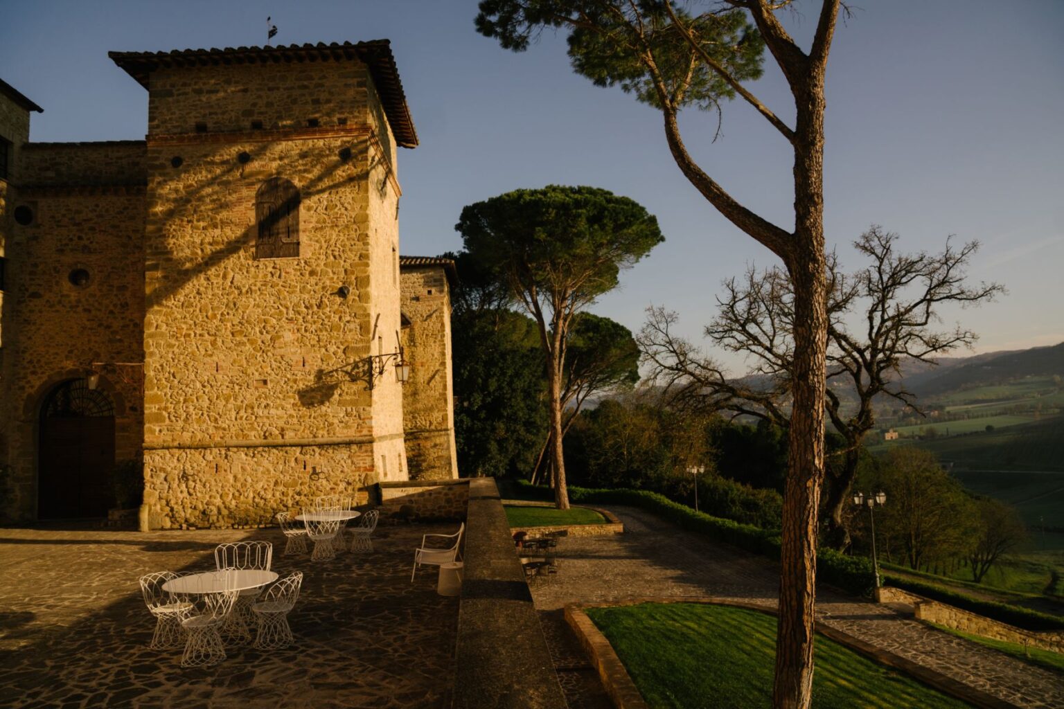 Historic stone building with a tower, surrounded by lush greenery and outdoor seating in a picturesque landscape. The scene captures the charm of an Italian villa at sunset, showcasing the architectural details and natural surroundings. | Only on SopranoVillas