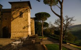 Historic stone building with a tower, surrounded by lush greenery and outdoor seating in a picturesque landscape. The scene captures the charm of an Italian villa at sunset, showcasing the architectural details and natural surroundings.
