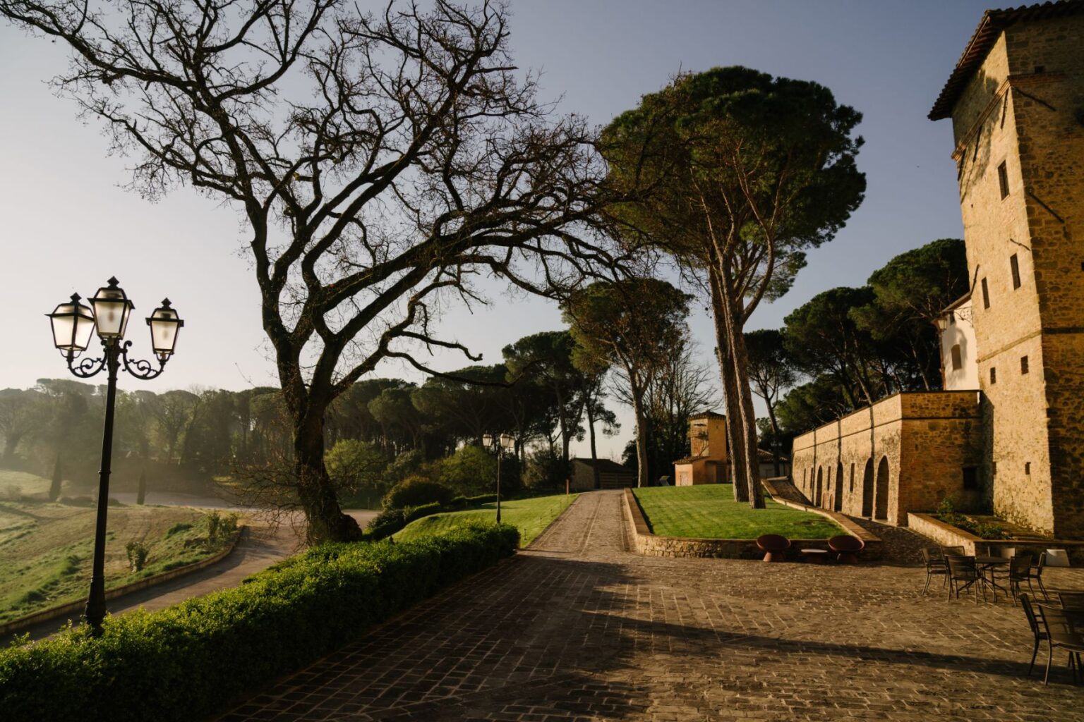 Scenic view of a historic stone building surrounded by lush greenery and tall trees, featuring a cobblestone pathway and vintage street lamps, set against a clear sky. Ideal for showcasing tranquil outdoor spaces in architectural photography. | Only on SopranoVillas