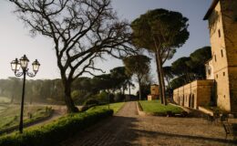 Scenic view of a historic stone building surrounded by lush greenery and tall trees, featuring a cobblestone pathway and vintage street lamps, set against a clear sky. Ideal for showcasing tranquil outdoor spaces in architectural photography.