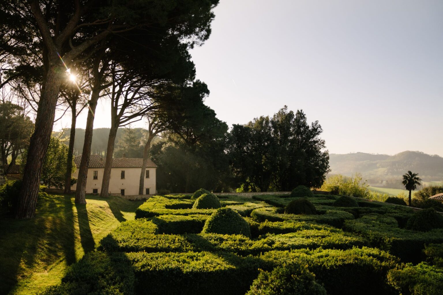 Scenic view of a landscaped garden with neatly trimmed hedges, a charming rustic house in the background, and tall trees casting shadows, set against a backdrop of rolling hills under a clear sky. Ideal for nature lovers and landscape photography enthusiasts. | Only on SopranoVillas
