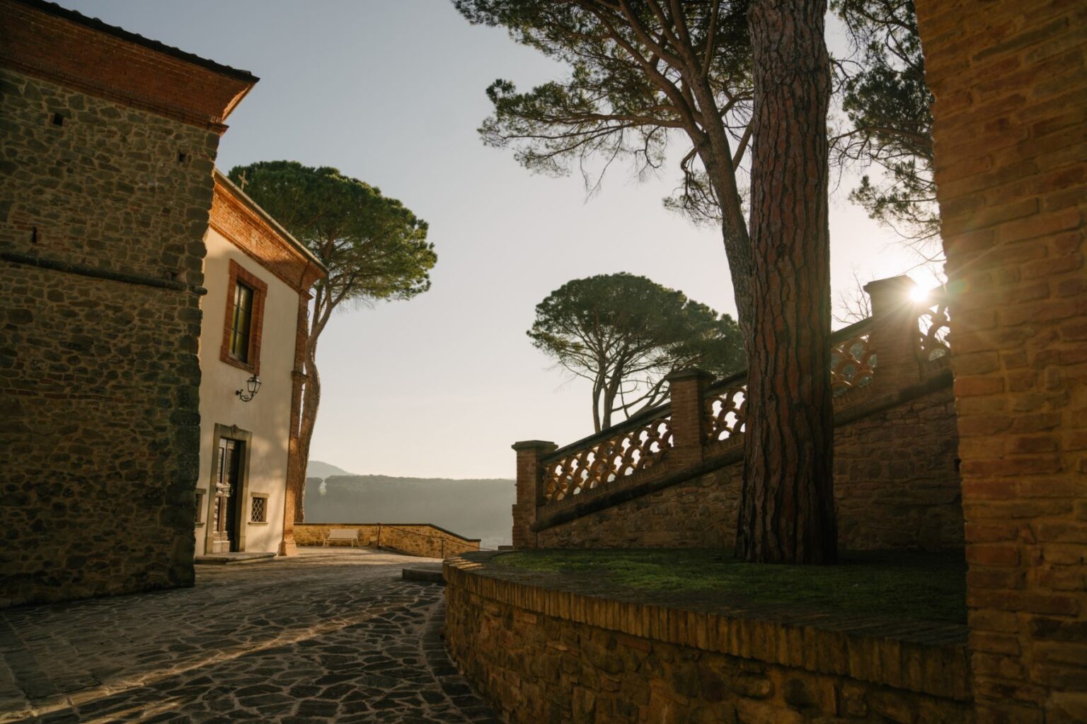 Scenic view of a historic stone building with a tree, featuring a cobblestone path and decorative railing, illuminated by soft morning light. | Only on SopranoVillas