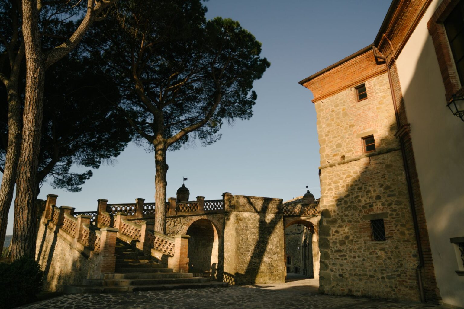 Historic stone architecture with a staircase leading to an arched gateway, surrounded by tall trees and a clear blue sky. The image captures the charm of Italian countryside buildings, featuring intricate stonework and a peaceful atmosphere. | Only on SopranoVillas