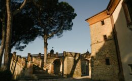 Historic stone architecture with a staircase leading to an arched gateway, surrounded by tall trees and a clear blue sky. The image captures the charm of Italian countryside buildings, featuring intricate stonework and a peaceful atmosphere.