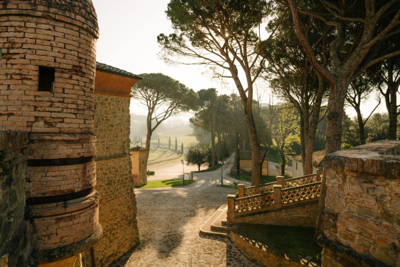 Scenic view of a historic stone building with a round tower, surrounded by tall trees and a winding pathway leading through a picturesque landscape in the early morning light. | Only on SopranoVillas