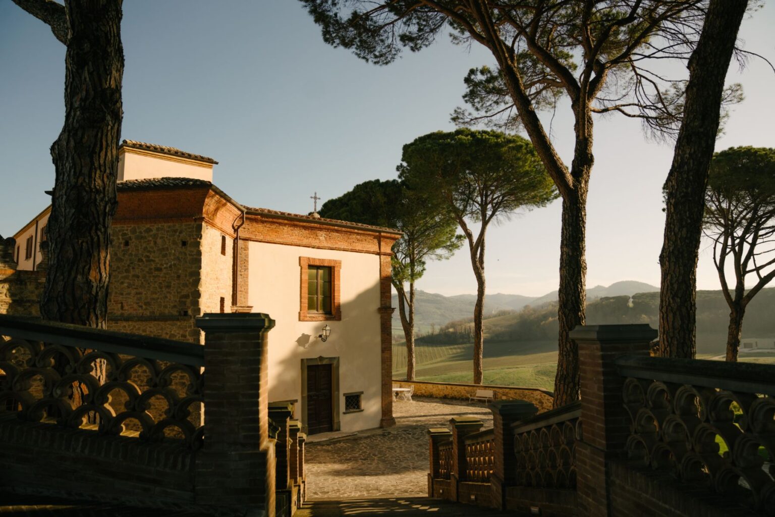 Scenic view of a rustic villa surrounded by tall trees, with a stone pathway leading to the entrance. The backdrop features rolling hills under a clear sky, highlighting the serene landscape of the countryside. | Only on SopranoVillas