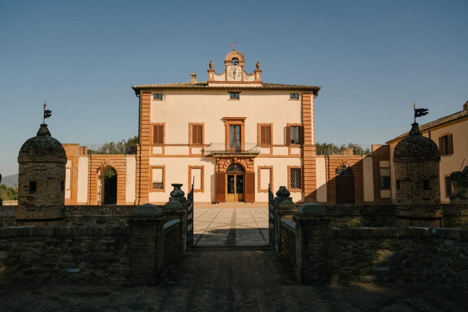 Historic Italian villa with distinctive architectural features, including a clock tower and decorative stonework, set against a clear blue sky. The entrance is framed by ornate gates and flanked by stone towers, showcasing the villa's elegant facade and surrounding landscape. | Only on SopranoVillas