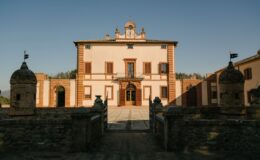 Historic Italian villa with distinctive architectural features, including a clock tower and decorative stonework, set against a clear blue sky. The entrance is framed by ornate gates and flanked by stone towers, showcasing the villa's elegant facade and surrounding landscape.