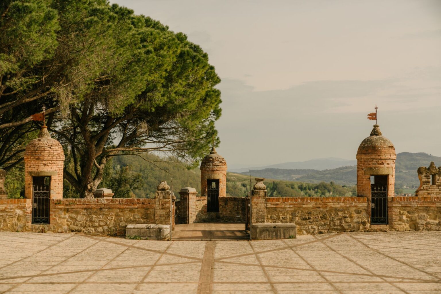 Historic stone wall with turrets and wrought iron gates, surrounded by lush greenery and mountains in the background, depicting a scenic view of an ancient fortress. | Only on SopranoVillas