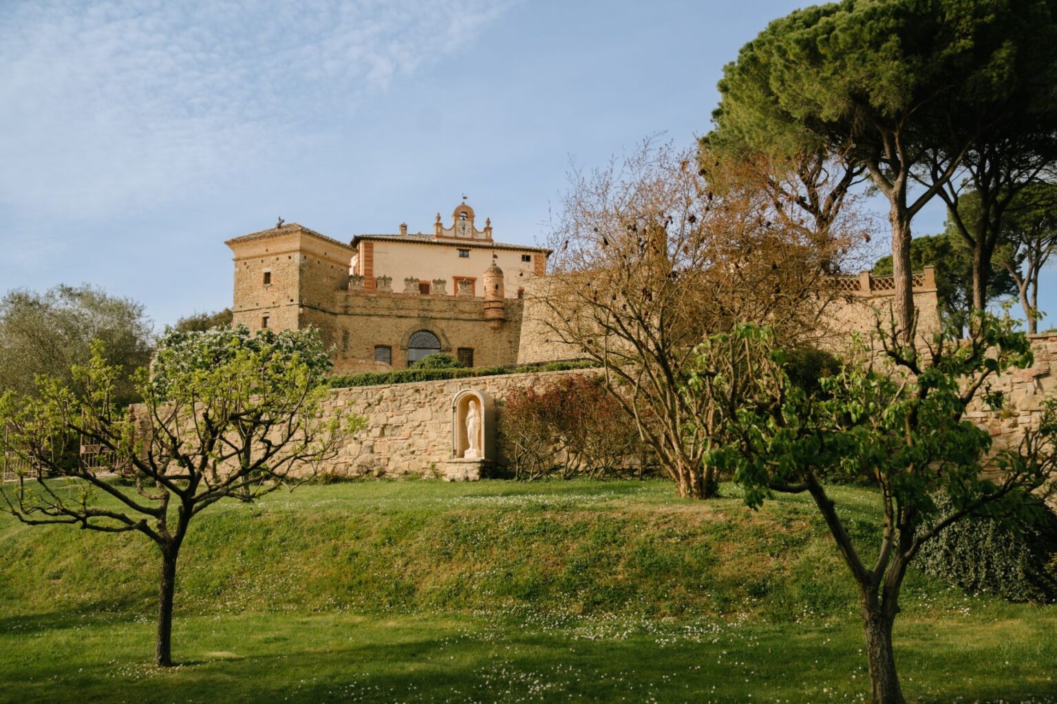 Historic villa surrounded by lush greenery and trees, showcasing a stone wall and a statue in the foreground, with a clear blue sky in the background. | Only on SopranoVillas