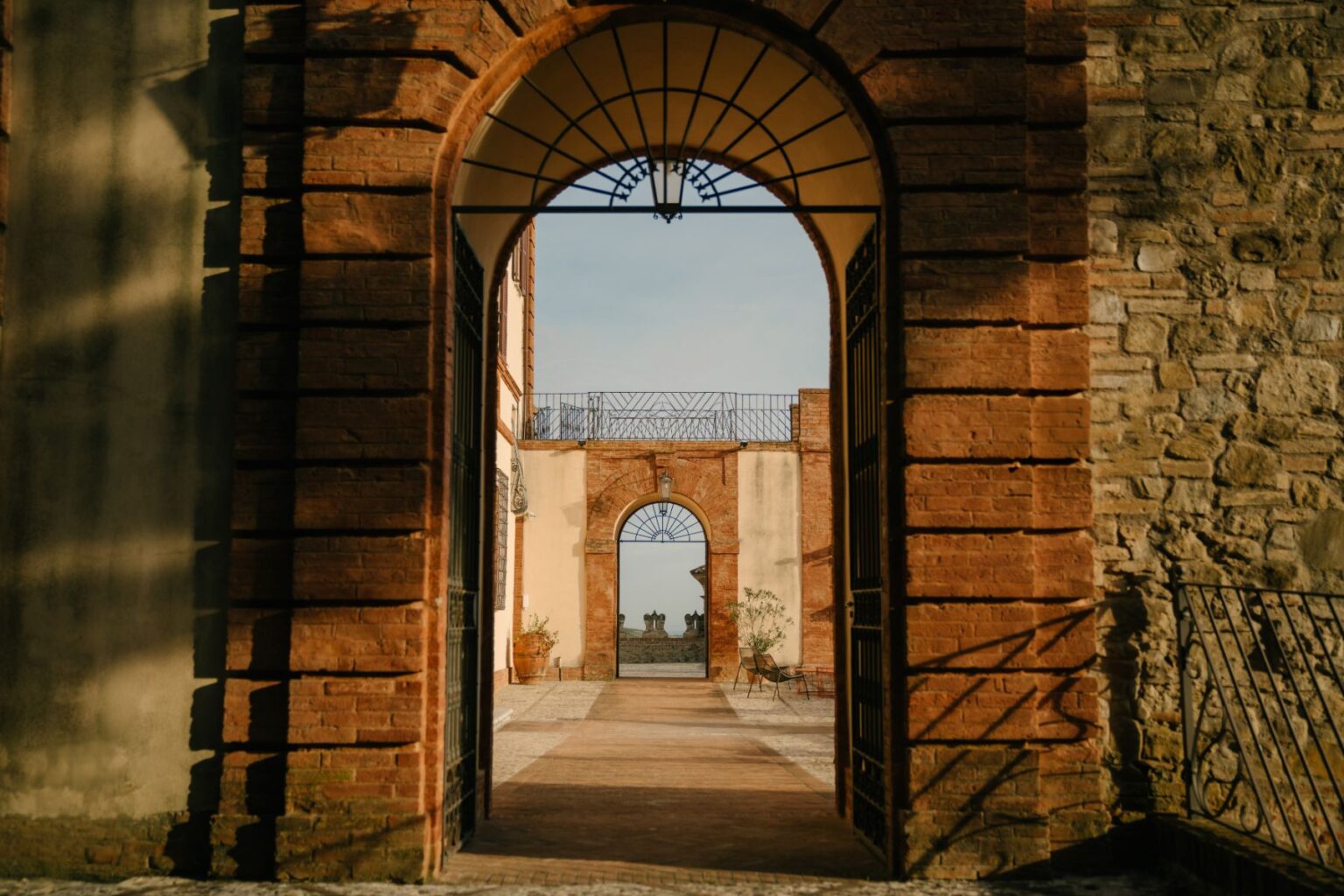 Archway view showcasing a historic building with brick and stone walls, leading to a courtyard with potted plants and a distant architectural feature under a clear sky. | Only on SopranoVillas