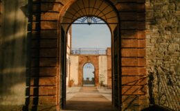 Archway view showcasing a historic building with brick and stone walls, leading to a courtyard with potted plants and a distant architectural feature under a clear sky.