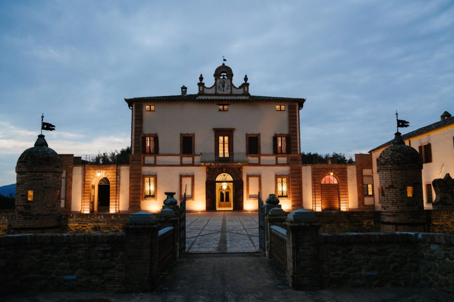 Elegant historic villa illuminated at dusk, featuring a grand entrance, arched windows, and stone towers. Ideal for luxury events and romantic getaways, set against a scenic landscape. | Only on SopranoVillas