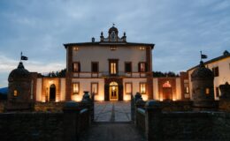 Elegant historic villa illuminated at dusk, featuring a grand entrance, arched windows, and stone towers. Ideal for luxury events and romantic getaways, set against a scenic landscape.