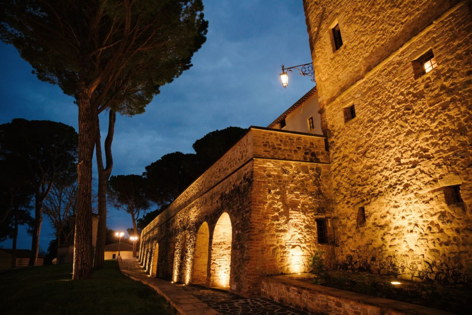 Historic stone building illuminated at night, featuring arched windows and a cobblestone path lined with trees and lamps. Ideal for showcasing architectural beauty and evening ambiance in outdoor settings. | Only on SopranoVillas