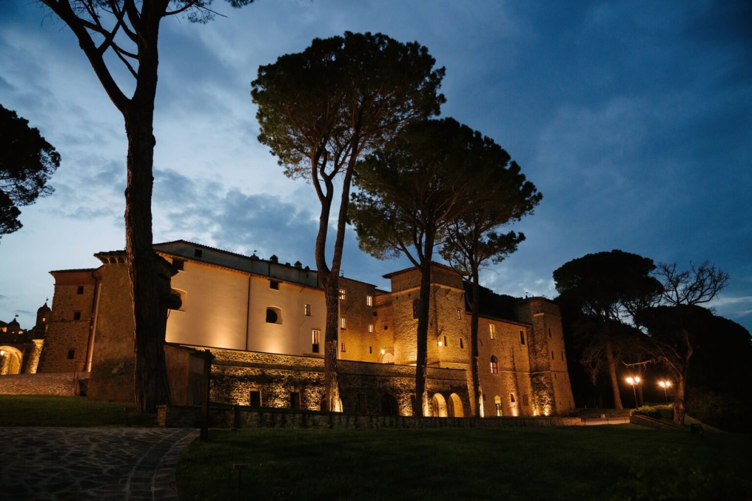 Historic stone castle illuminated at dusk, surrounded by tall pine trees and a serene landscape, showcasing architectural details and a peaceful atmosphere. | Only on SopranoVillas