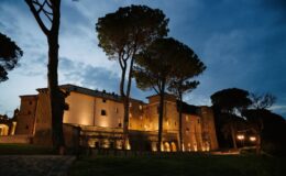 Historic stone castle illuminated at dusk, surrounded by tall pine trees and a serene landscape, showcasing architectural details and a peaceful atmosphere.
