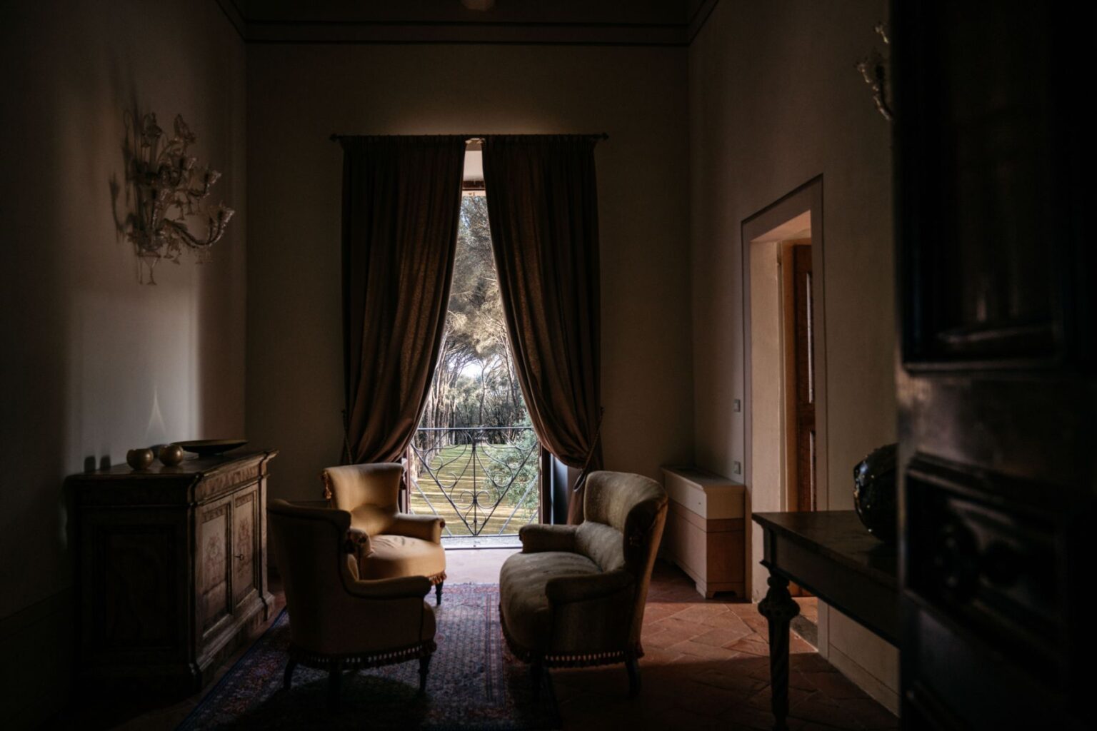 Cozy living room with two elegant armchairs and a vintage wooden cabinet, featuring large curtains framing a view of greenery through a balcony door, soft natural light illuminating the space. | Only on SopranoVillas