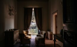 Cozy living room with two elegant armchairs and a vintage wooden cabinet, featuring large curtains framing a view of greenery through a balcony door, soft natural light illuminating the space.