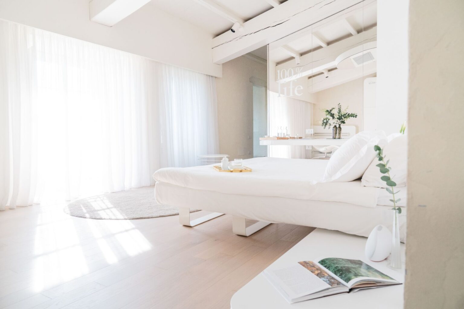 Bright and airy bedroom with white curtains, a minimalist bed, and a cozy rug, featuring a small table with a decorative vase and an open magazine, emphasizing modern design and tranquility. | Only on SopranoVillas