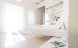 Bright and airy bedroom with white curtains, a minimalist bed, and a cozy rug, featuring a small table with a decorative vase and an open magazine, emphasizing modern design and tranquility.