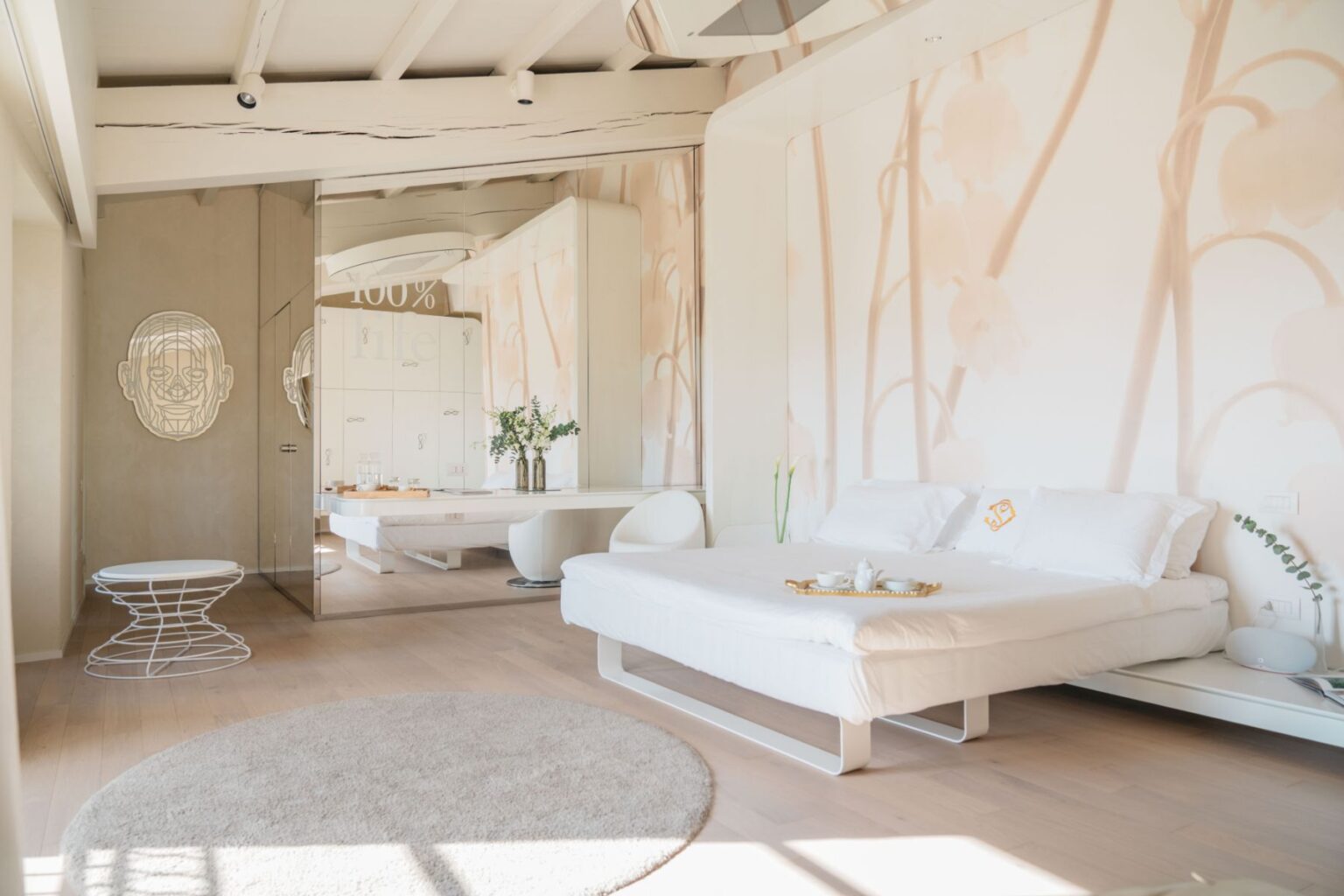 Modern bedroom interior featuring a minimalist design with a white bed, soft rug, and elegant decor. The walls are adorned with light floral patterns, and a unique mirrored wall reflects a modern art piece. A small table holds a tea set, and natural light floods the space, creating a serene atmosphere. | Only on SopranoVillas
