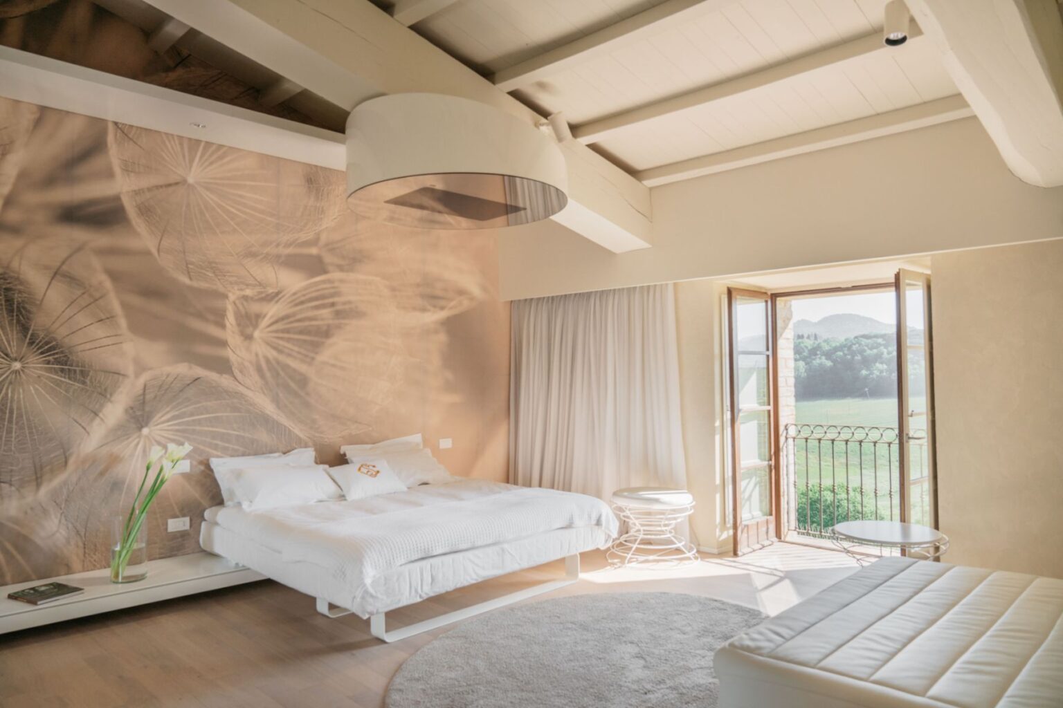 Bright and airy bedroom featuring a large bed with white linens, minimalist furniture, and a scenic view through open French doors. The room is adorned with a nature-themed wall mural and has wooden beams and a soft area rug for added comfort. Ideal for relaxation and tranquility. | Only on SopranoVillas