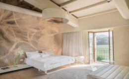 Bright and airy bedroom featuring a large bed with white linens, minimalist furniture, and a scenic view through open French doors. The room is adorned with a nature-themed wall mural and has wooden beams and a soft area rug for added comfort. Ideal for relaxation and tranquility.