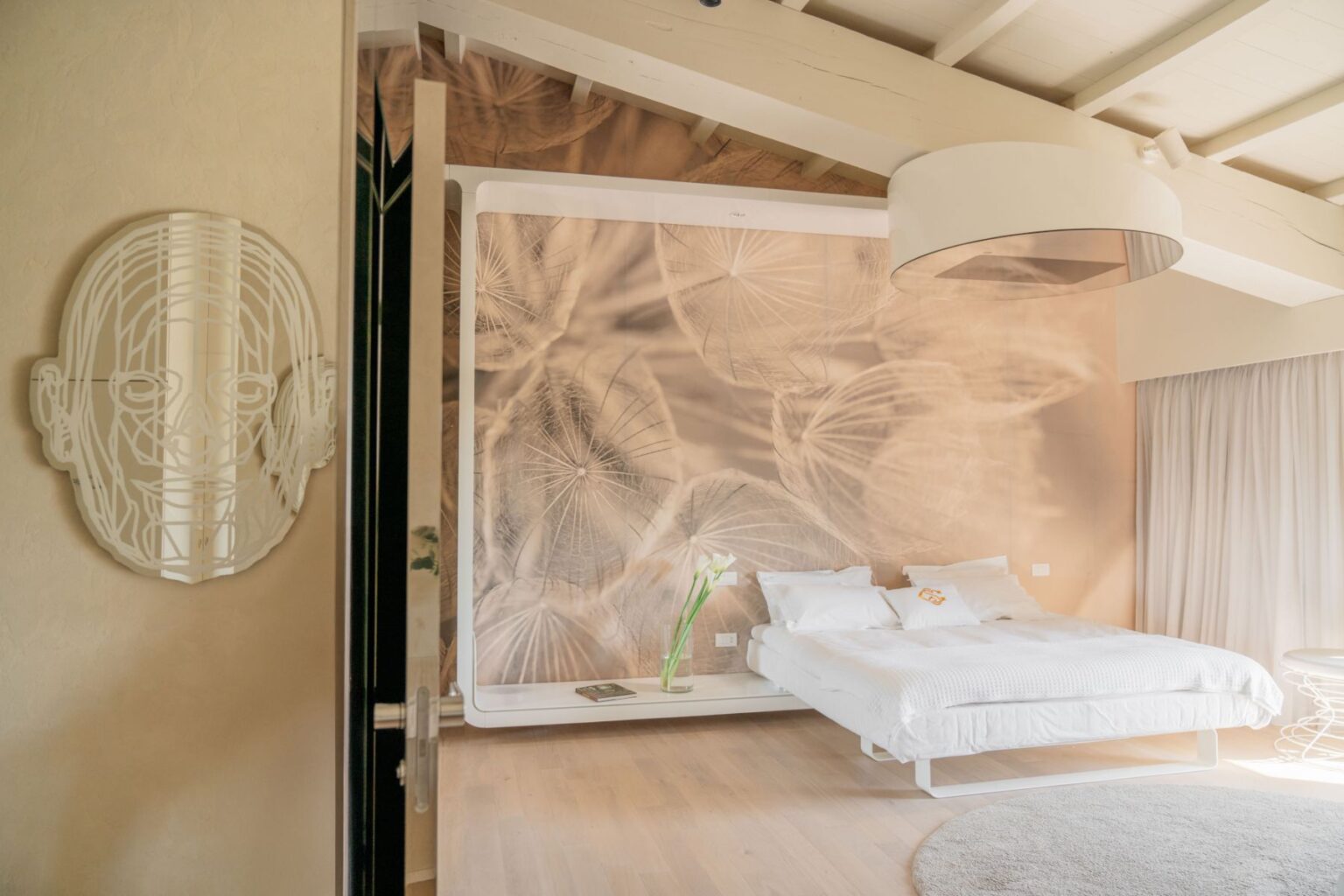 Modern bedroom interior featuring a white bed with crisp linens, a decorative wall mural of delicate plant life, and a unique wall art piece resembling a face. The room has natural light, wooden flooring, and minimalistic decor for a serene atmosphere. | Only on SopranoVillas
