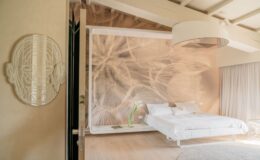 Modern bedroom interior featuring a white bed with crisp linens, a decorative wall mural of delicate plant life, and a unique wall art piece resembling a face. The room has natural light, wooden flooring, and minimalistic decor for a serene atmosphere.
