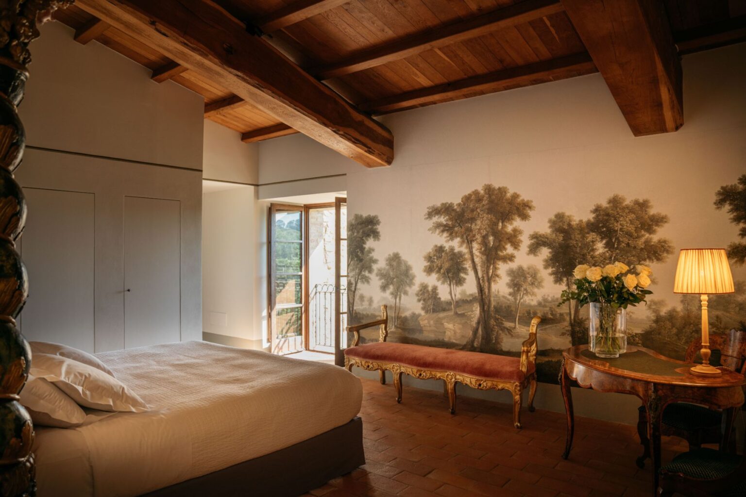 Cozy bedroom featuring a plush bed with white linens, a vintage-style bench, and a decorative wooden table with a vase of white roses. Natural light floods the room through an open window, highlighting a scenic mural of trees on the wall and wooden beam ceiling, creating a warm and inviting atmosphere. | Only on SopranoVillas