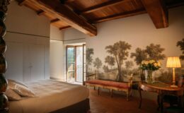 Cozy bedroom featuring a plush bed with white linens, a vintage-style bench, and a decorative wooden table with a vase of white roses. Natural light floods the room through an open window, highlighting a scenic mural of trees on the wall and wooden beam ceiling, creating a warm and inviting atmosphere.
