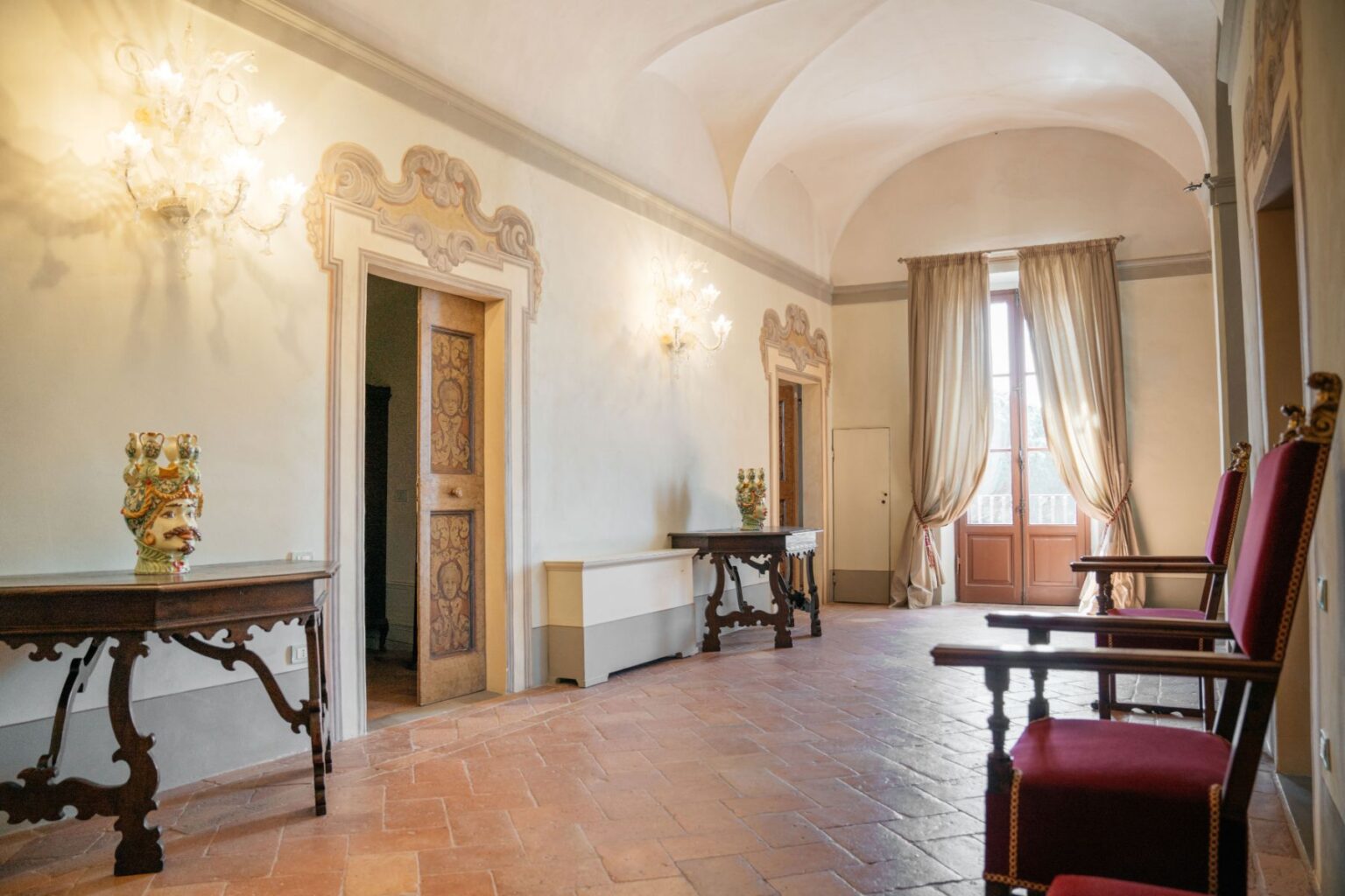 Elegant interior of a historic room featuring arched ceilings, vintage furniture, and ornate decor. The space includes a decorative chandelier, a stylish table with a ceramic centerpiece, and plush red chairs, enhancing the room's classic charm. Natural light streams in through large windows, creating a warm and inviting atmosphere. | Only on SopranoVillas