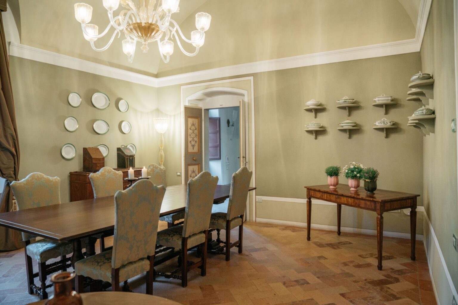 Elegant dining room featuring a long wooden table with upholstered chairs, adorned with decorative plates on the walls, a vintage side cabinet, and a small table with potted plants. The room is illuminated by a stylish chandelier and has a soothing green color palette. | Only on SopranoVillas