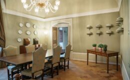 Elegant dining room featuring a long wooden table with upholstered chairs, adorned with decorative plates on the walls, a vintage side cabinet, and a small table with potted plants. The room is illuminated by a stylish chandelier and has a soothing green color palette.