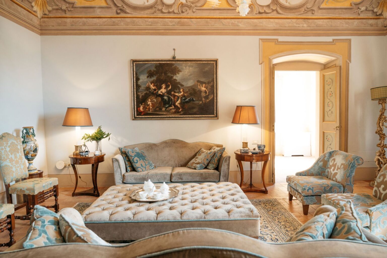 Elegant living room featuring plush sofas with decorative cushions, a tufted coffee table, and classic artwork on the walls. The space is enhanced by stylish lamps and ornate furniture, creating a sophisticated atmosphere. Natural light streams in through an open door, illuminating the intricate ceiling details. | Only on SopranoVillas
