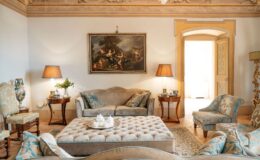 Elegant living room featuring plush sofas with decorative cushions, a tufted coffee table, and classic artwork on the walls. The space is enhanced by stylish lamps and ornate furniture, creating a sophisticated atmosphere. Natural light streams in through an open door, illuminating the intricate ceiling details.