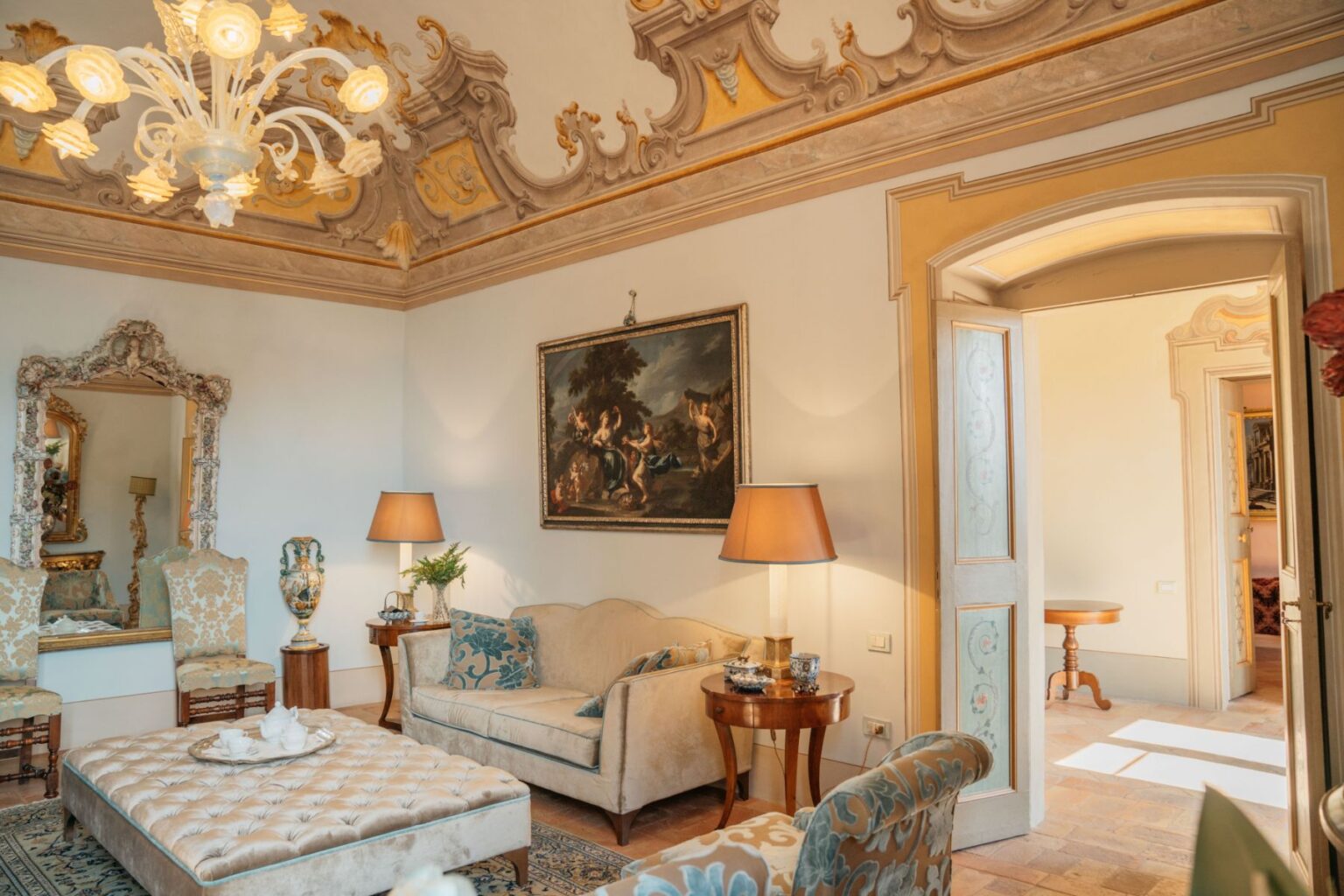 Elegant living room featuring ornate ceiling details, vintage furniture, and a classic chandelier. The space includes a plush sofa, decorative armchairs, a coffee table with tea service, and a large mirror, creating a warm and inviting atmosphere. | Only on SopranoVillas