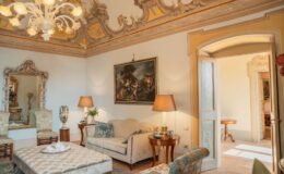 Elegant living room featuring ornate ceiling details, vintage furniture, and a classic chandelier. The space includes a plush sofa, decorative armchairs, a coffee table with tea service, and a large mirror, creating a warm and inviting atmosphere.