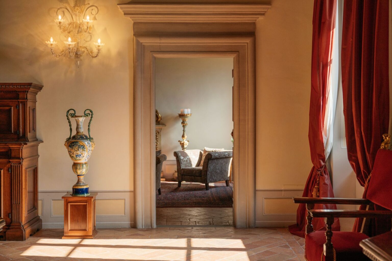 Elegant interior view showcasing a decorative vase on a pedestal, ornate chandelier, and a cozy sitting area visible through an archway. The room features warm lighting, rich textures, and classic furnishings, creating a sophisticated ambiance. | Only on SopranoVillas