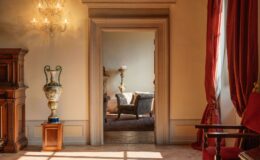 Elegant interior view showcasing a decorative vase on a pedestal, ornate chandelier, and a cozy sitting area visible through an archway. The room features warm lighting, rich textures, and classic furnishings, creating a sophisticated ambiance.