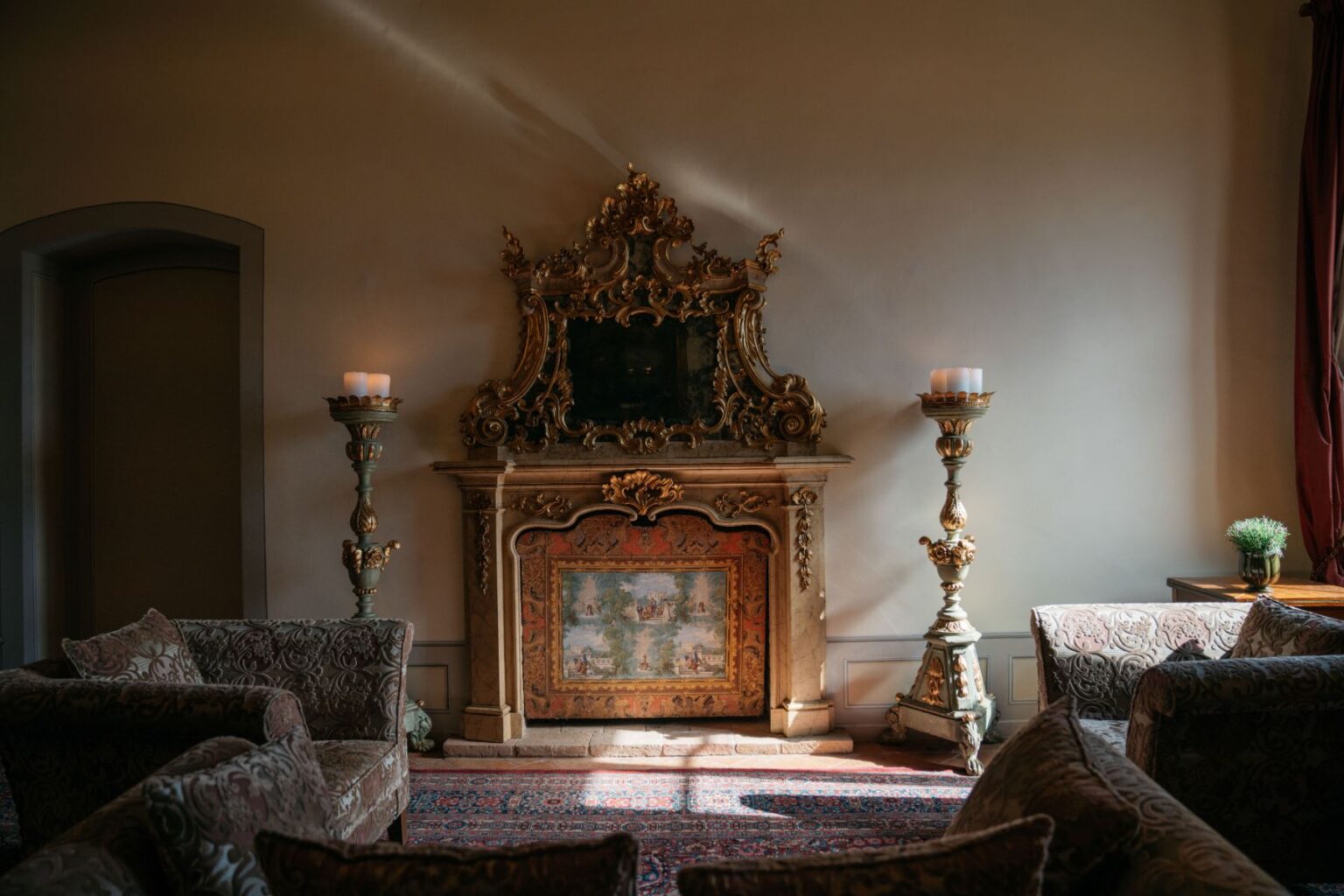 Elegant living room featuring an ornate fireplace with a decorative mirror, flanked by tall candle holders. Plush sofas with intricate patterns and a vibrant area rug complete the sophisticated interior design. Soft natural light enhances the warm ambiance. | Only on SopranoVillas