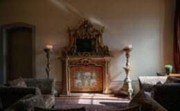 Elegant living room featuring an ornate fireplace with a decorative mirror, flanked by tall candle holders. Plush sofas with intricate patterns and a vibrant area rug complete the sophisticated interior design. Soft natural light enhances the warm ambiance.