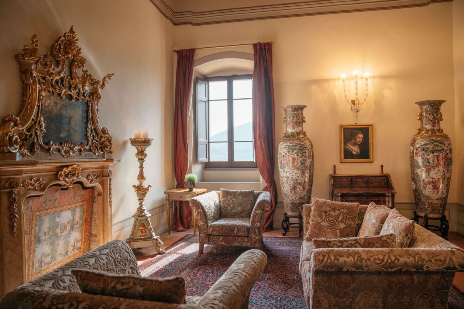 Luxurious living room featuring ornate furniture, a grand fireplace with intricate carvings, two large decorative vases, elegant drapes, and a window offering a scenic view. The space is adorned with a painting and a small side table with a potted plant, creating a sophisticated ambiance. | Only on SopranoVillas