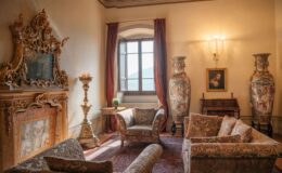 Luxurious living room featuring ornate furniture, a grand fireplace with intricate carvings, two large decorative vases, elegant drapes, and a window offering a scenic view. The space is adorned with a painting and a small side table with a potted plant, creating a sophisticated ambiance.