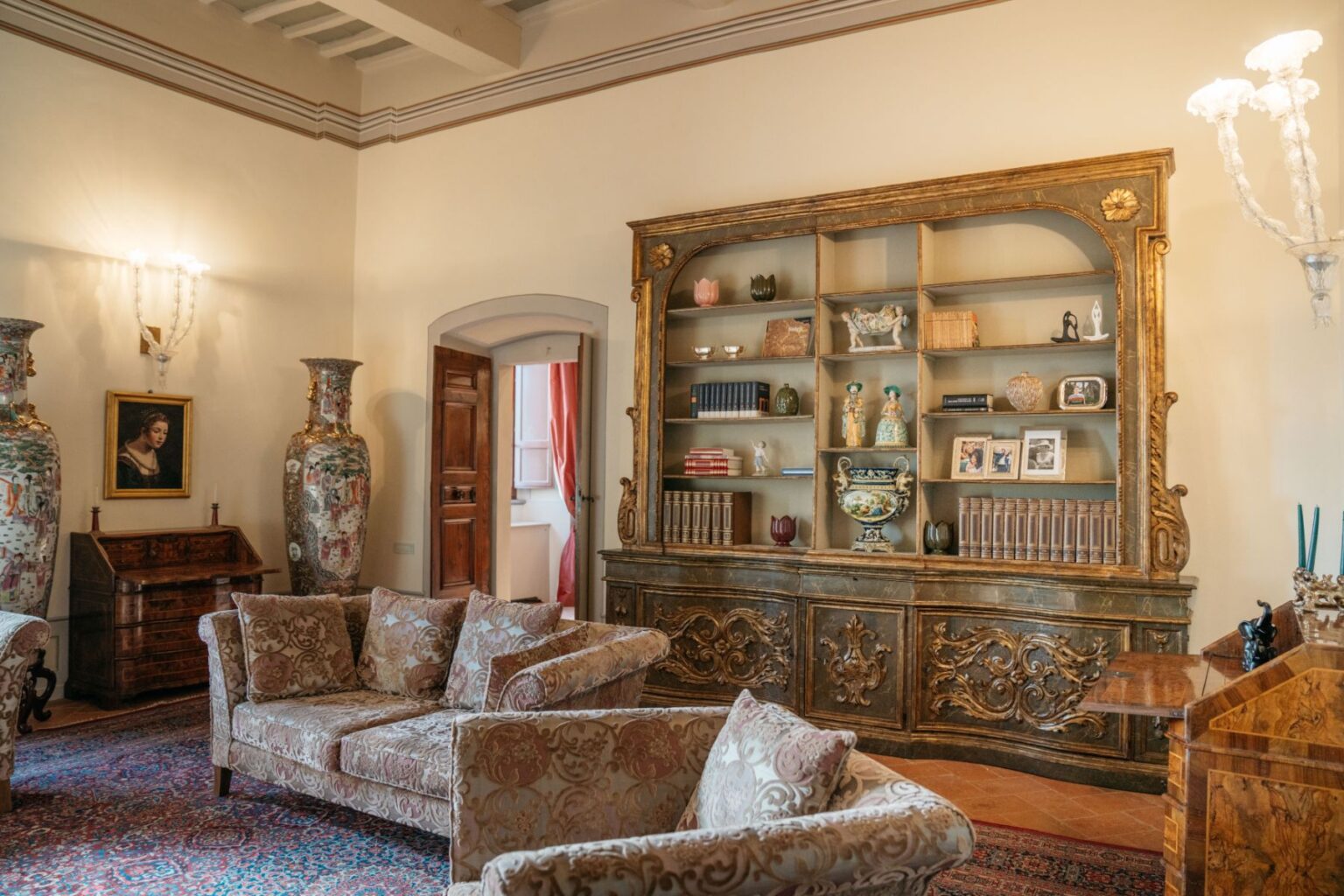 Elegant living room featuring ornate furniture, including plush sofas and a decorative bookshelf filled with books and collectibles. Two large vases stand prominently, complemented by a vintage writing desk and a painting on the wall. The room showcases classic design elements with warm lighting and soft color tones. | Only on SopranoVillas