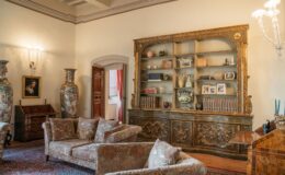 Elegant living room featuring ornate furniture, including plush sofas and a decorative bookshelf filled with books and collectibles. Two large vases stand prominently, complemented by a vintage writing desk and a painting on the wall. The room showcases classic design elements with warm lighting and soft color tones.